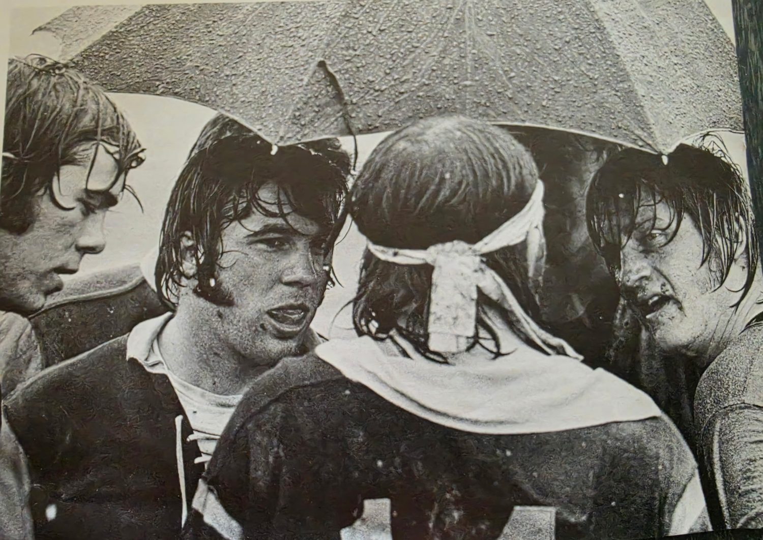 Four soaked Mizzou players crowded under an umbrella in the rain, one wearing a scrum cap tied at the back