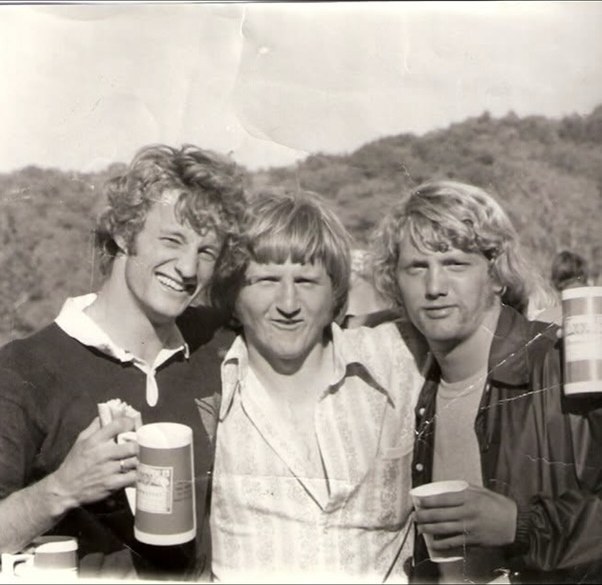 Three young Mizzou ruggers with beer steins after a match, wooded hills behind them