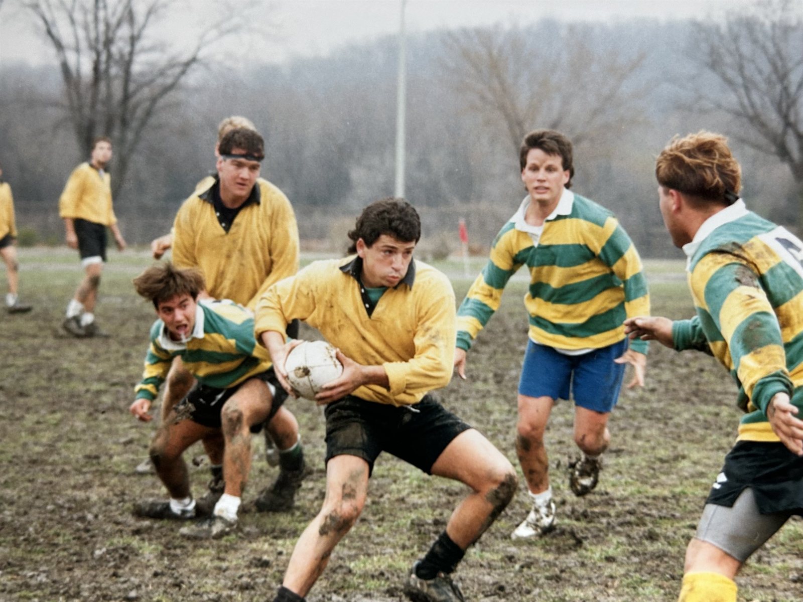 A Mizzou ball carrier in a gold jersey breaks through a green-and-gold hooped defense on a muddy field during the 1990-91 season