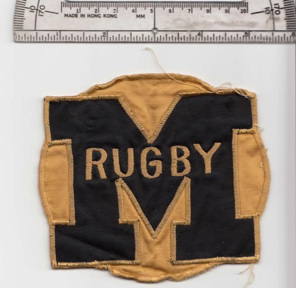 The original Mizzou Rugby patch: a black shield with a gold block M and the word RUGBY across it, frayed at the edges, photographed beside a ruler