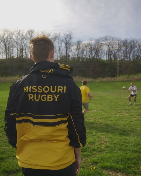 The back of a black and gold Missouri Rugby jacket with tiger-stripe detailing, worn on the touchline as players pass a ball behind