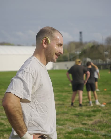 A Mizzou player in a white shirt stands smiling on the training pitch with teammates stretching in the background