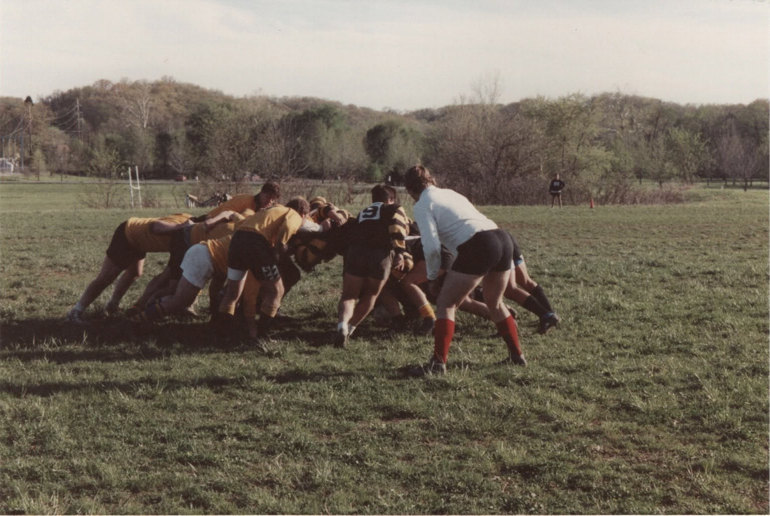 A scrum packs down on a bare pitch with a wooded hillside behind