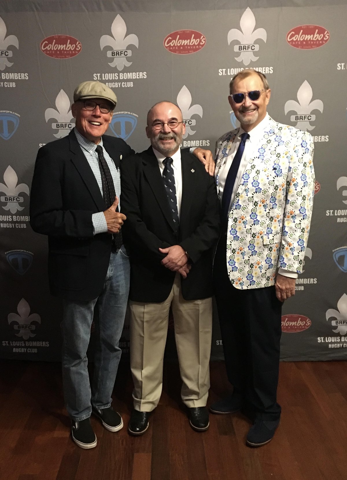 Three men in jackets and ties in front of a St. Louis Bombers Rugby Club backdrop, John Tarpoff in the center
