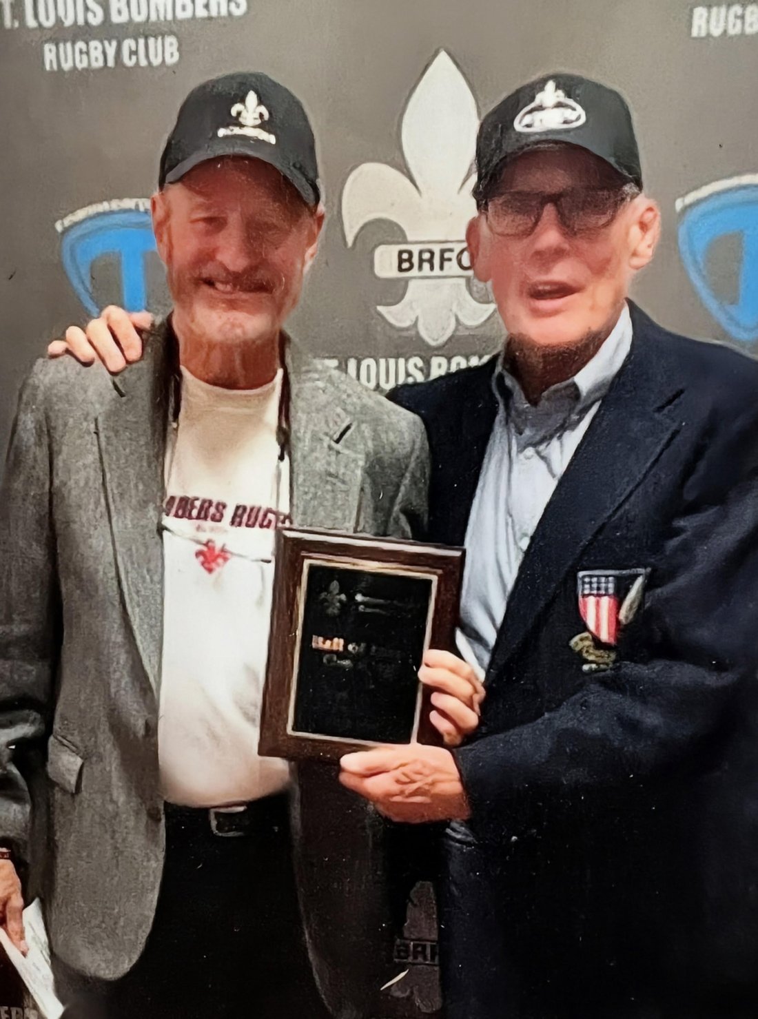 Rick Wetzel, left, and Jim Dierker in caps holding a Hall of Fame plaque between them in front of a St. Louis Bombers Rugby Club backdrop